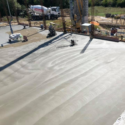 Blick auf einen Baustellenbereich mit frisch gegossenem Betonboden, Baumaschinen und einem Lastwagen im Hintergrund.